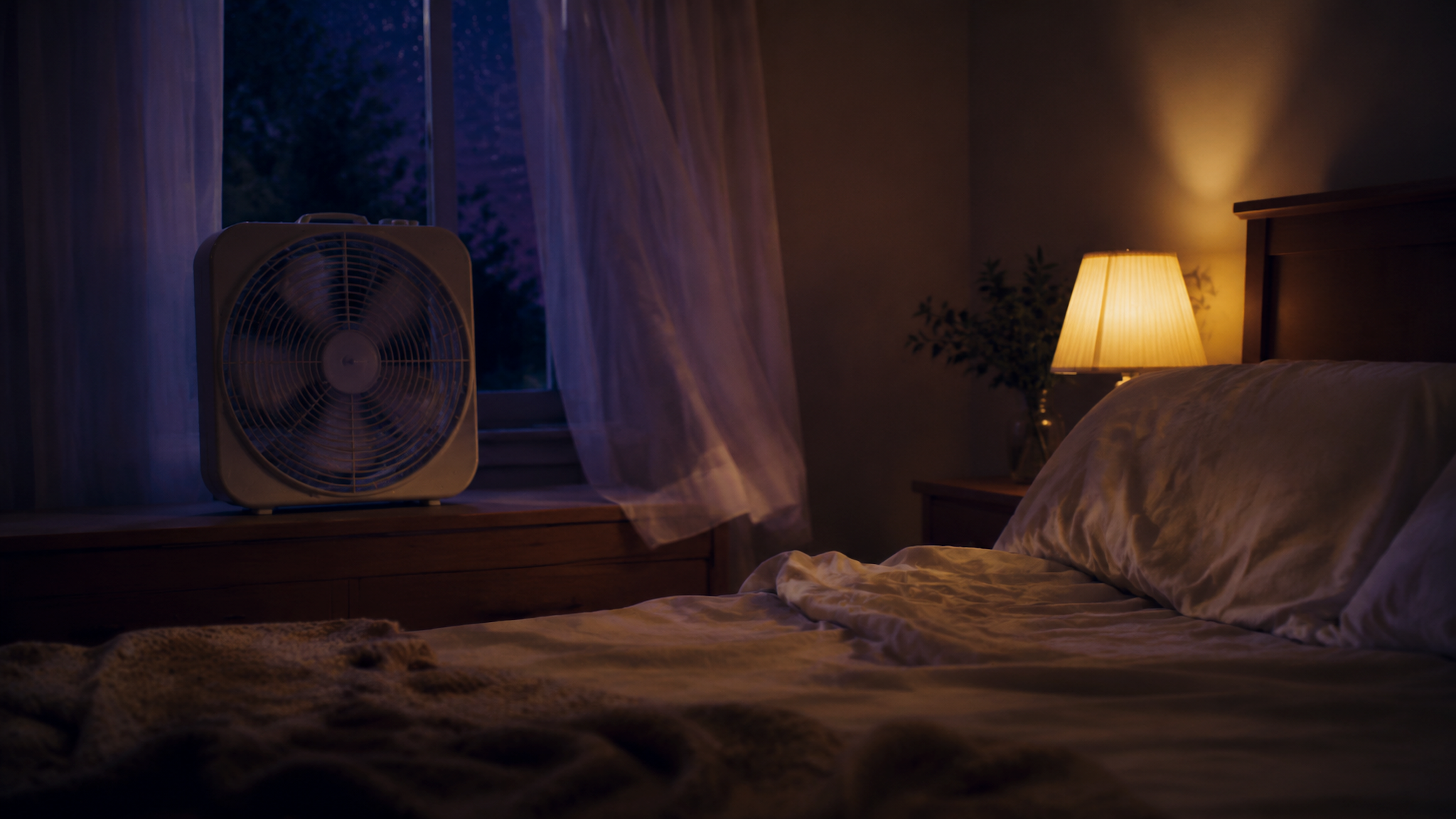A dim bedroom at night with a box fan in front of a window, sheer curtains drifting in its breeze, rumpled bedding, and a warm bedside lamp against a blue-purple sky.