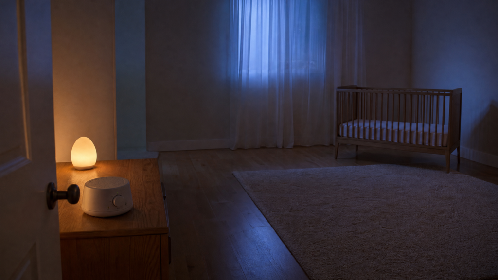 A dim nursery at night seen from the doorway, with a small cream sound machine and a warm night light on a dresser by the door, and an empty crib with only a fitted sheet far across the room under blue moonlight.